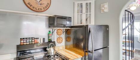Una cocina con estufa, microondas y refrigerador. Hay una tabla de cortar de madera, un armario y un reloj de pared.