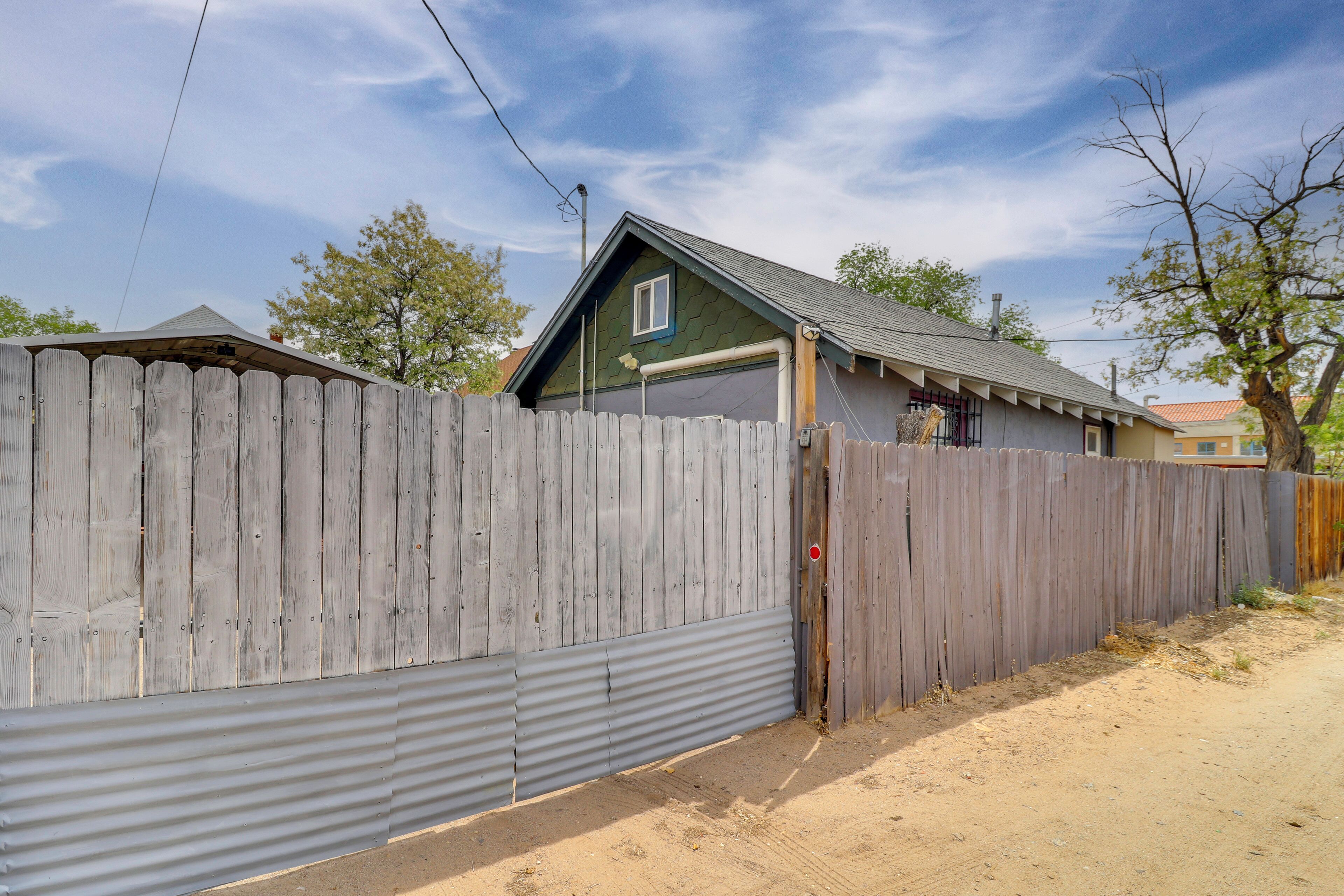 A fenced property with a corrugated metal wall, a wooden fence, and a house with a green roof and a window.