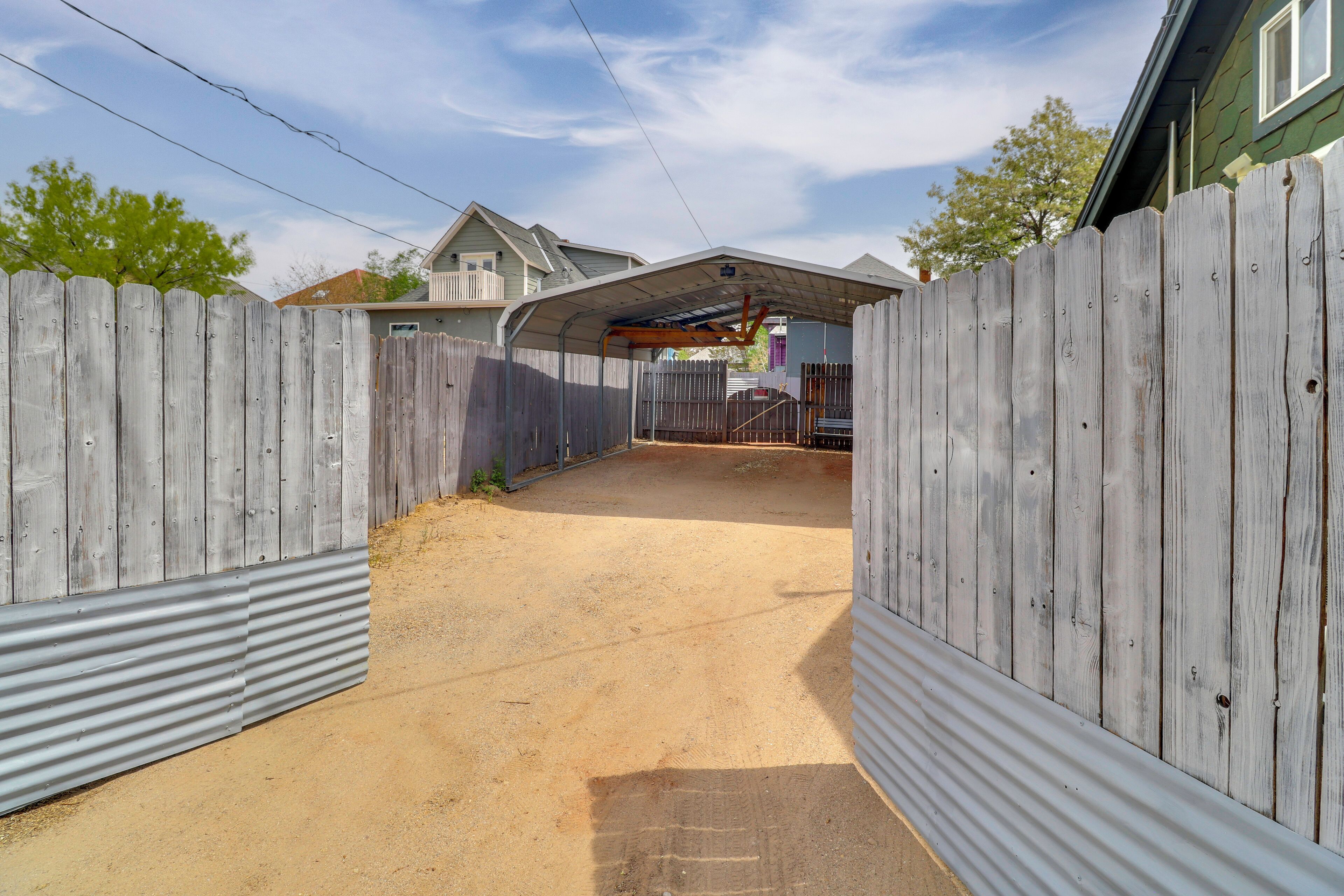 A fenced backyard with a corrugated metal wall, a covered area, and a house in the background.