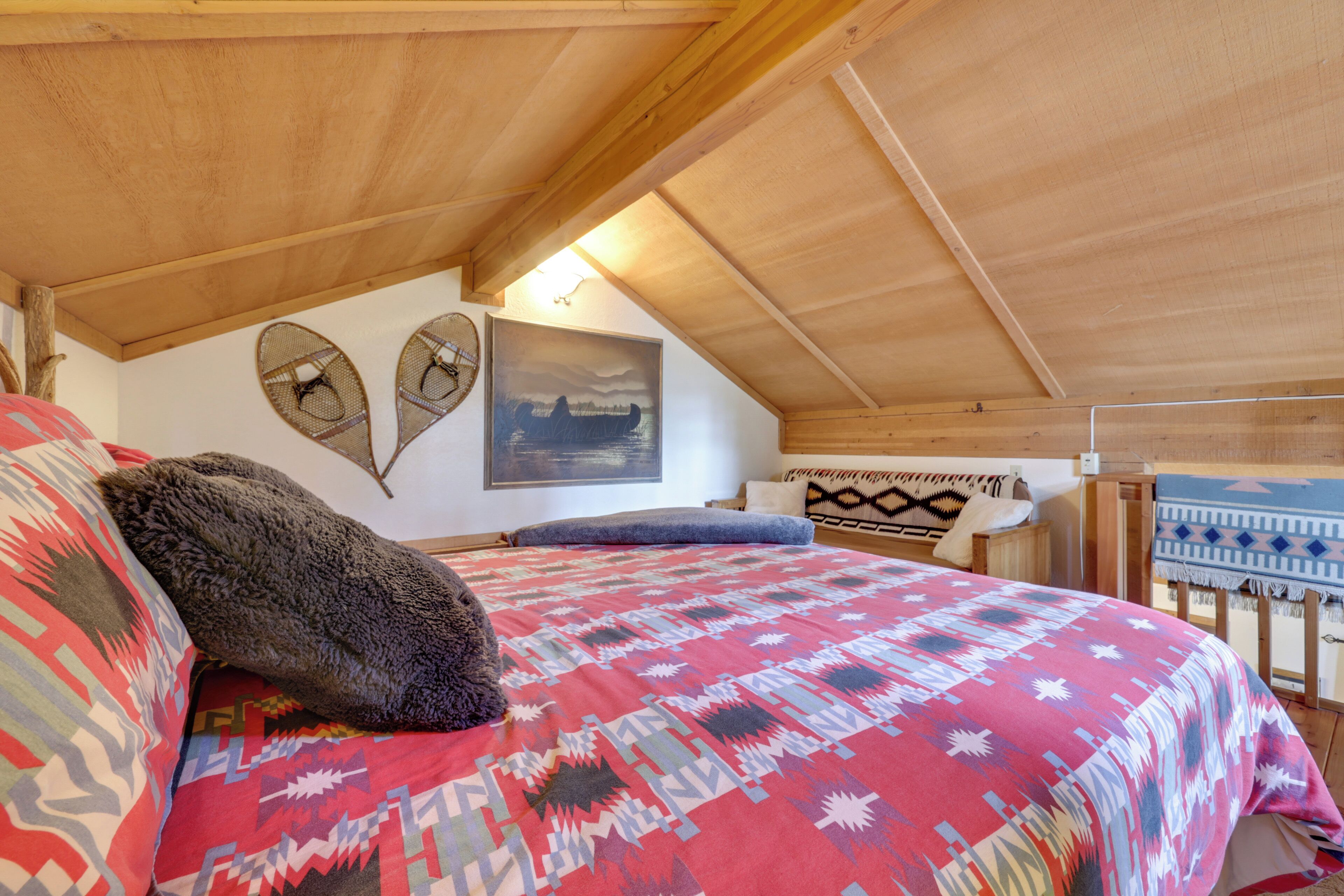 A bedroom with a wooden ceiling, a bed with a patterned blanket, two wall-mounted fans, and a painting.