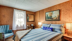 Un dormitorio con una cama, una silla, una mesita de noche, una lámpara, un cuadro y una ventana con cortinas.
