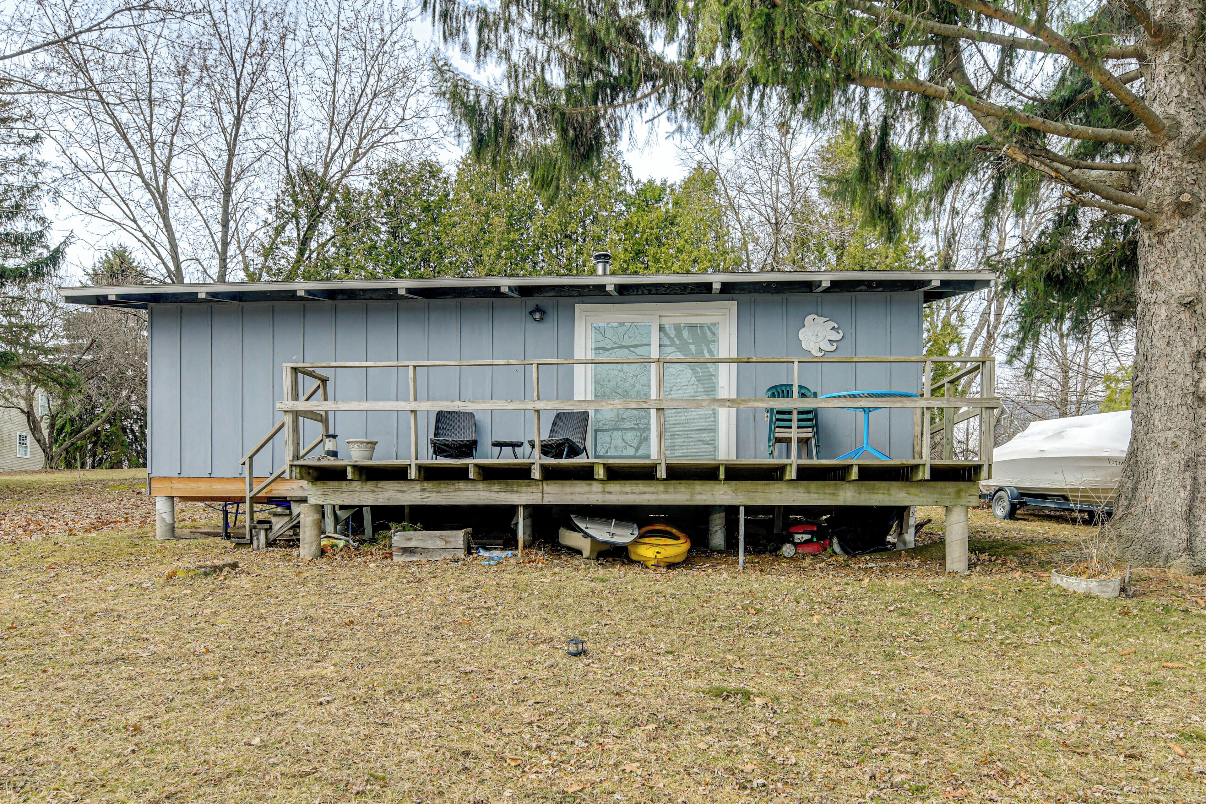 A small, single-story cabin with a deck, a covered area with chairs, and a boat in the background.
