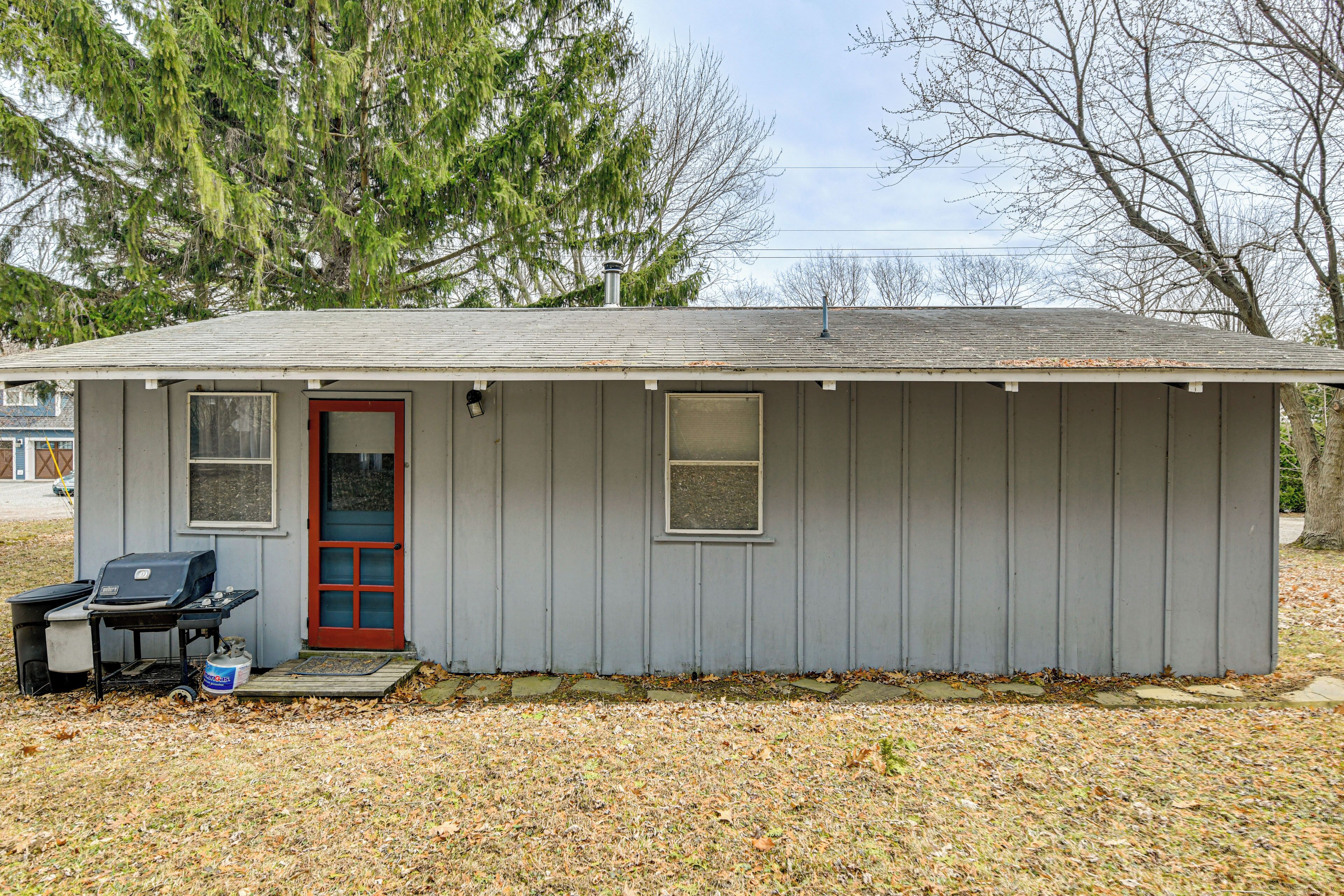 A small, single-story building with a red door, two windows, and a covered outdoor area with a grill.