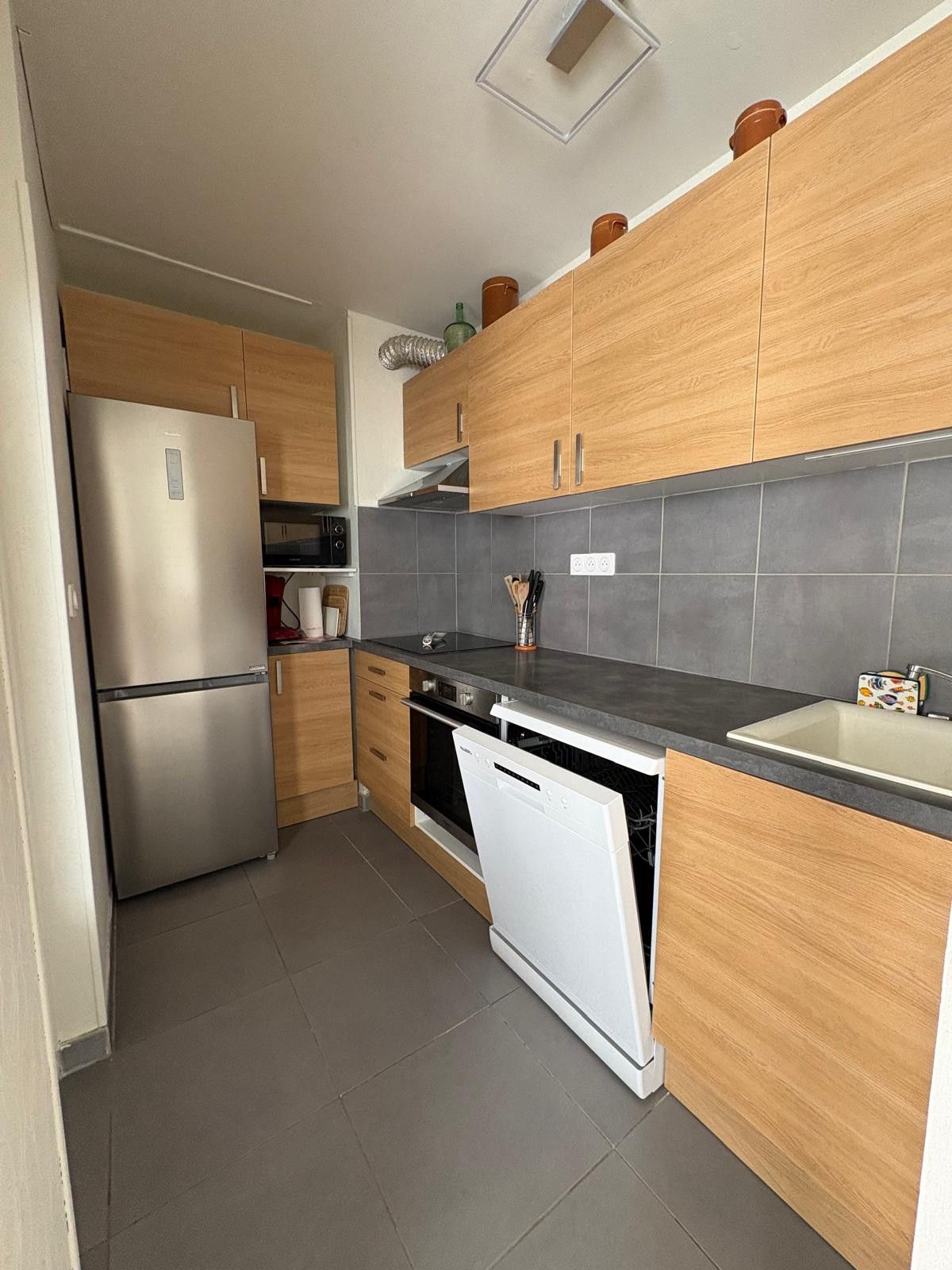 Cocina compacta con armarios de madera, un refrigerador de acero inoxidable, lavavajillas y salpicadero de azulejos grises.