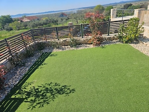 A fenced outdoor area with artificial grass, a stone wall, and a view of distant houses and landscape. - Holiday Home A&M with pool and lakeview (Vrana)