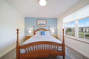 A bedroom with a four-poster bed, two bedside tables with lamps, a framed picture on the wall, and a large window with a view of buildings. - 421 - Sand-tastic Adventures Await - Oceanview (Westport)