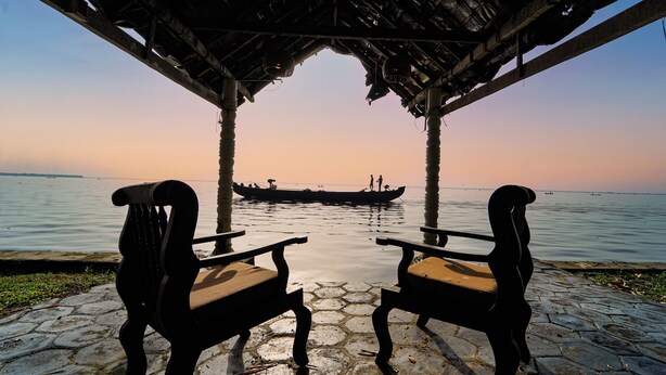 A stone-paved area with two wooden chairs facing a calm body of water. - Alaya Stays by ELIVAAS Vembanad House  (Cherthala)