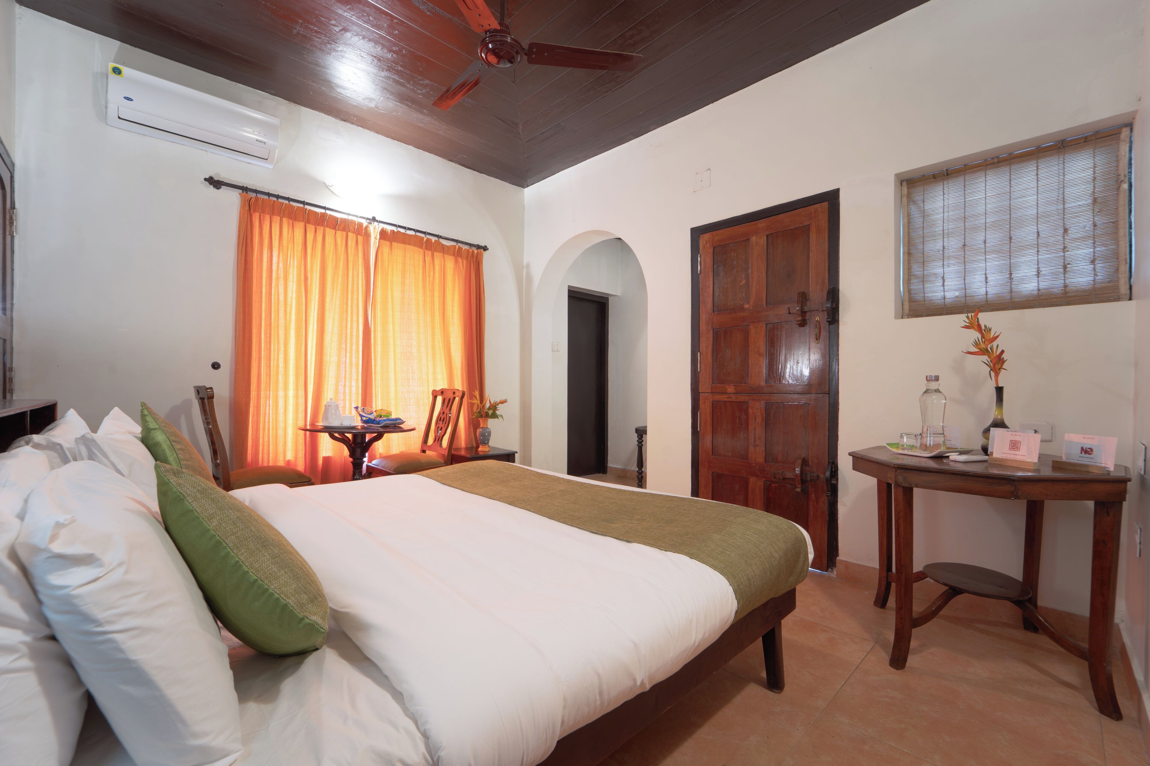A hotel room with a bed, an air conditioner, a ceiling fan, an arched doorway, a wooden door, a window with blinds, and a small table with a vase.