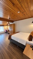 Habitación de hotel con techo de madera, cama con edredón blanco, escritorio con silla y ventana con cortinas.