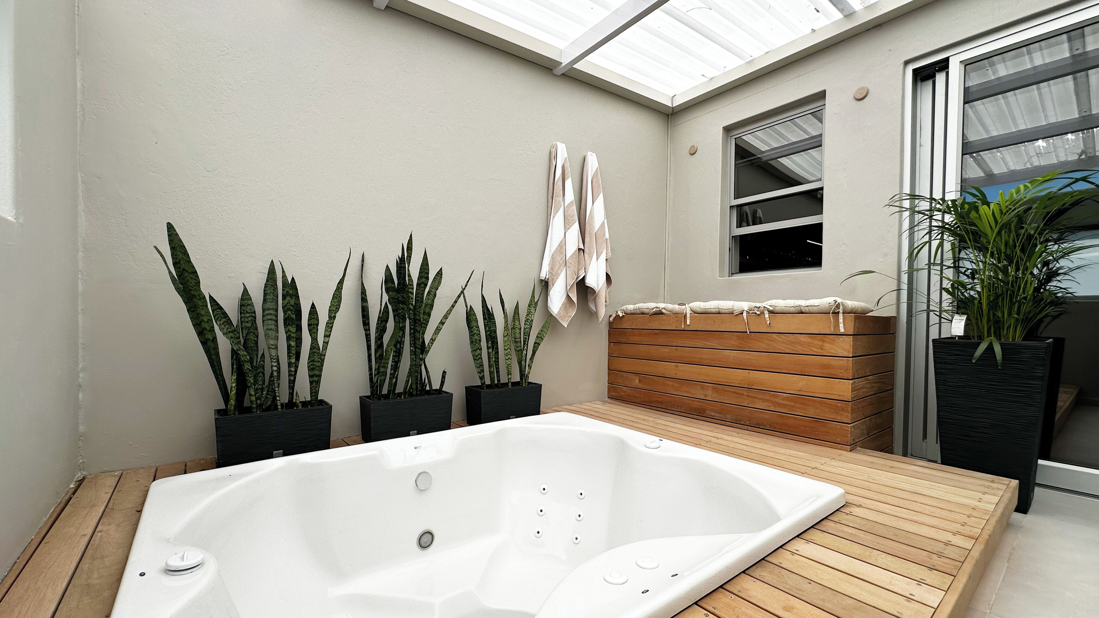 Een wellnessruimte met een jacuzzi, houten terras, plantenbakken en een raam.