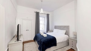 A bedroom with a bed, a bedside table, a lamp, a window with curtains, and a door. - Stylish West End 1-Bed | Prime Location (Edinburgh)