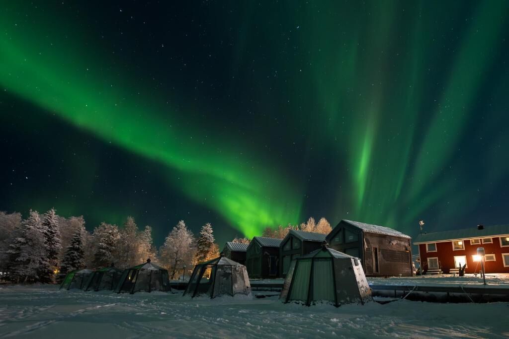 A snowy outdoor area with tents and a building, under a night sky with green aurora.