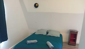 Een kleine, eenvoudige kamer met een bed, kussens en een nachtkastje.