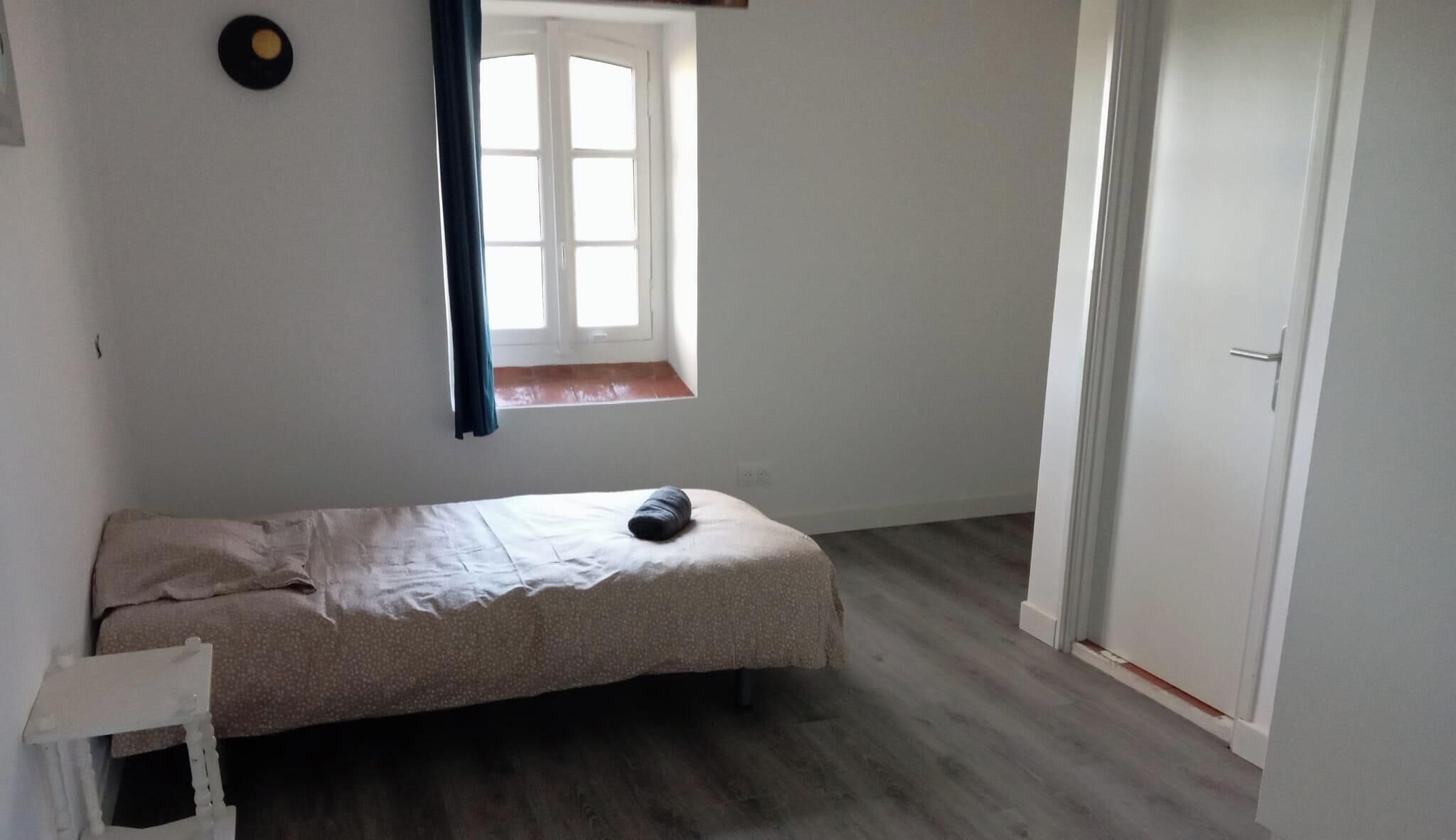 Een kleine, eenvoudig ingerichte kamer met een enkel bed, een raam met gordijnen, een deur en een kleine tafel.