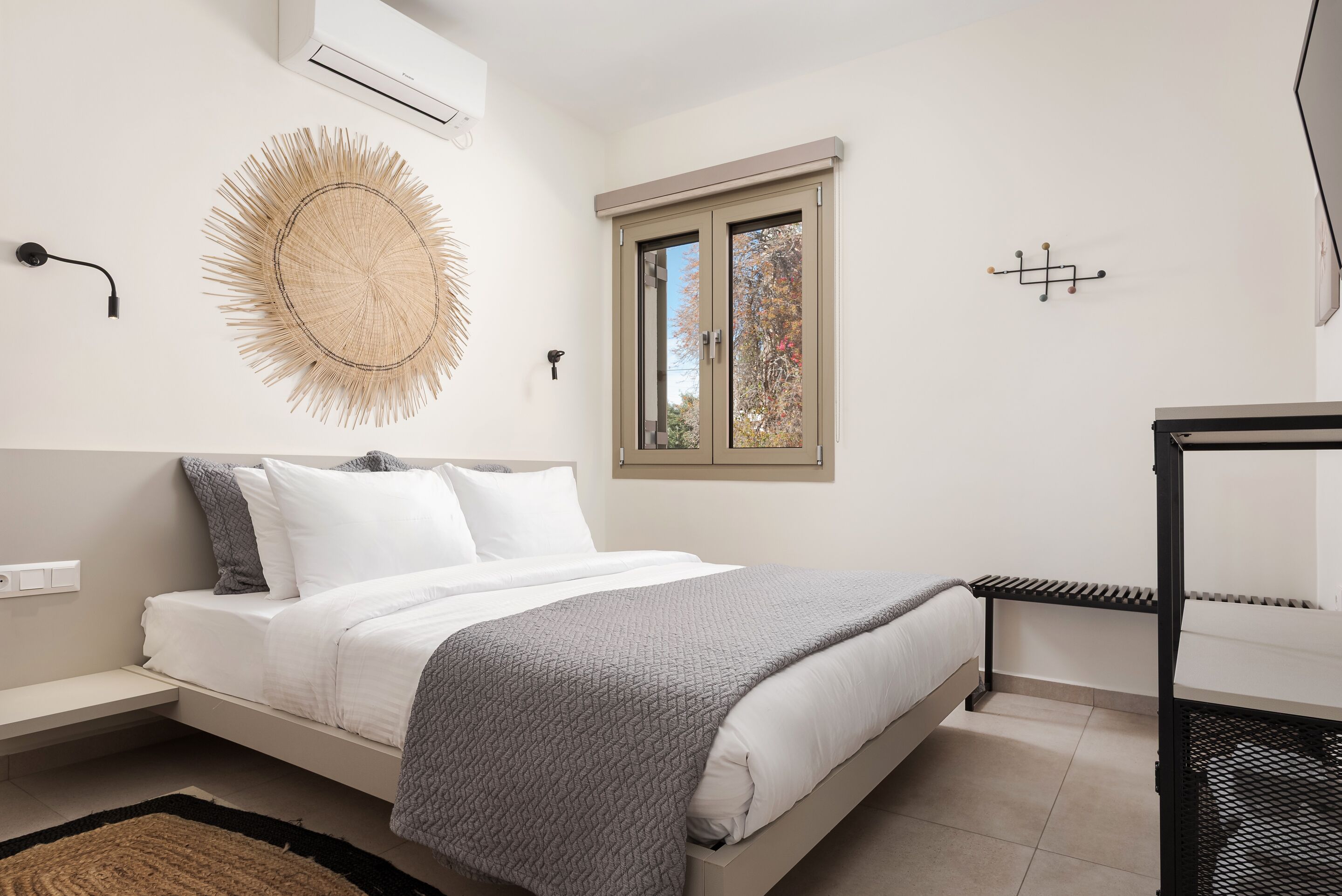 A modern bedroom with a bed, bedside table, wall-mounted air conditioning unit, decorative wall art, a window with a view, and a black metal bench.