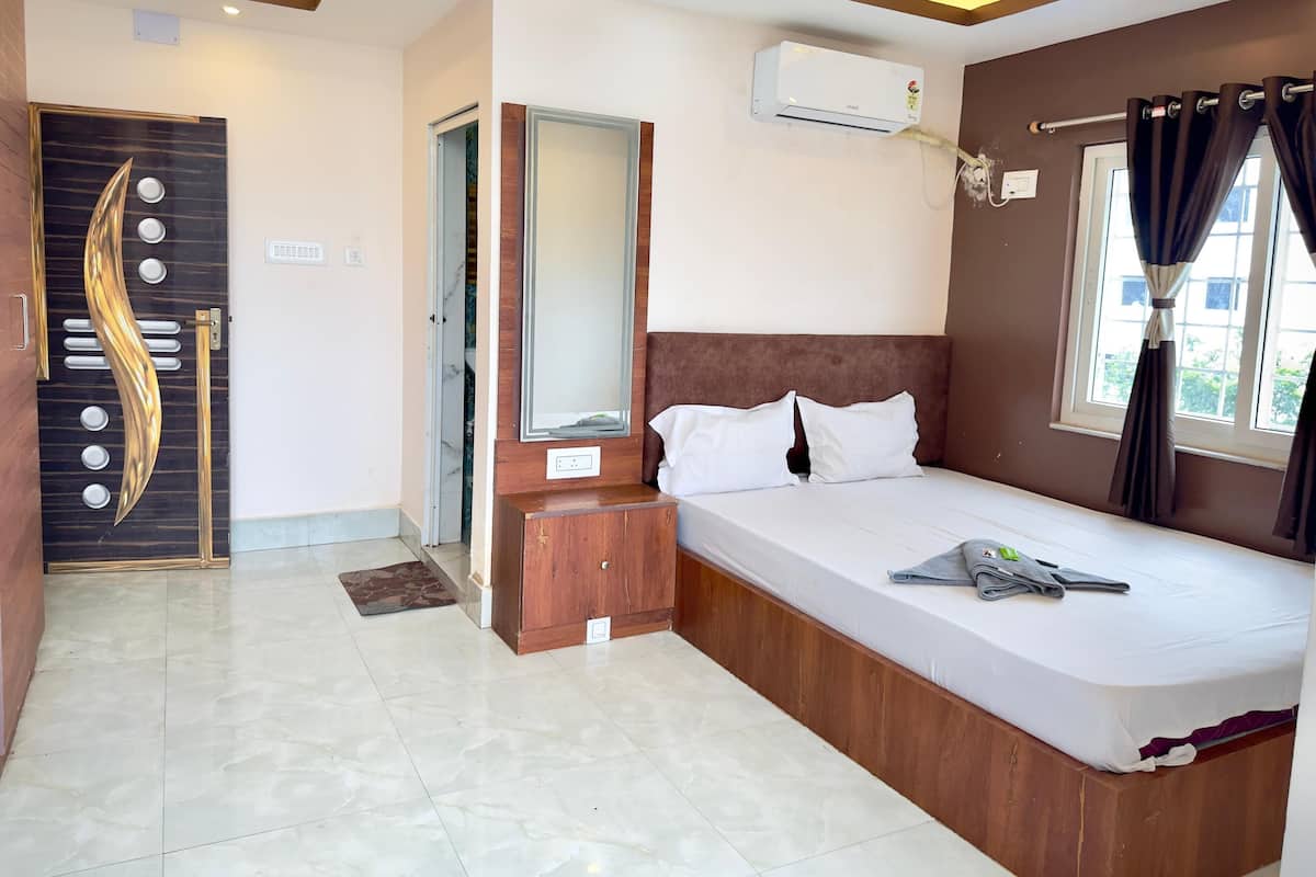 A hotel room with a bed, bedside table, mirror, air conditioning unit, and a window with curtains.