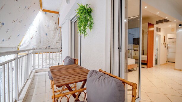 Een balkon met een houten tafel en rieten stoelen, een schuifdeur naar een slaapkamer en een hangplant.