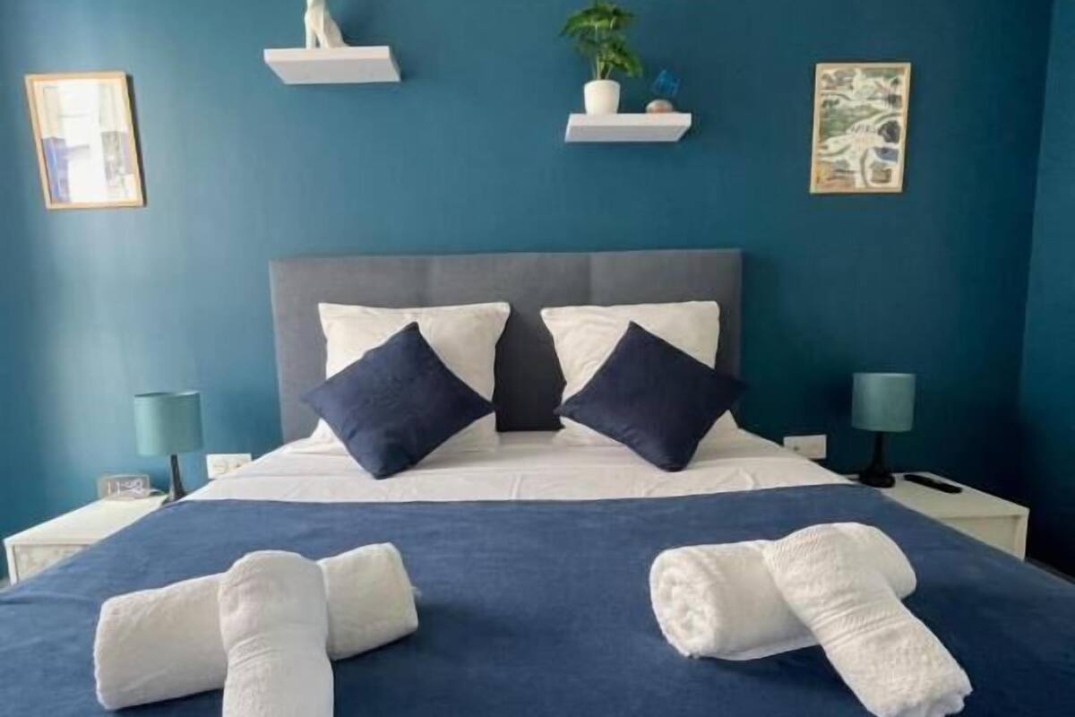 A neatly made bed with pillows, a nightstand with a lamp, and wall decorations including a plant and a framed picture.