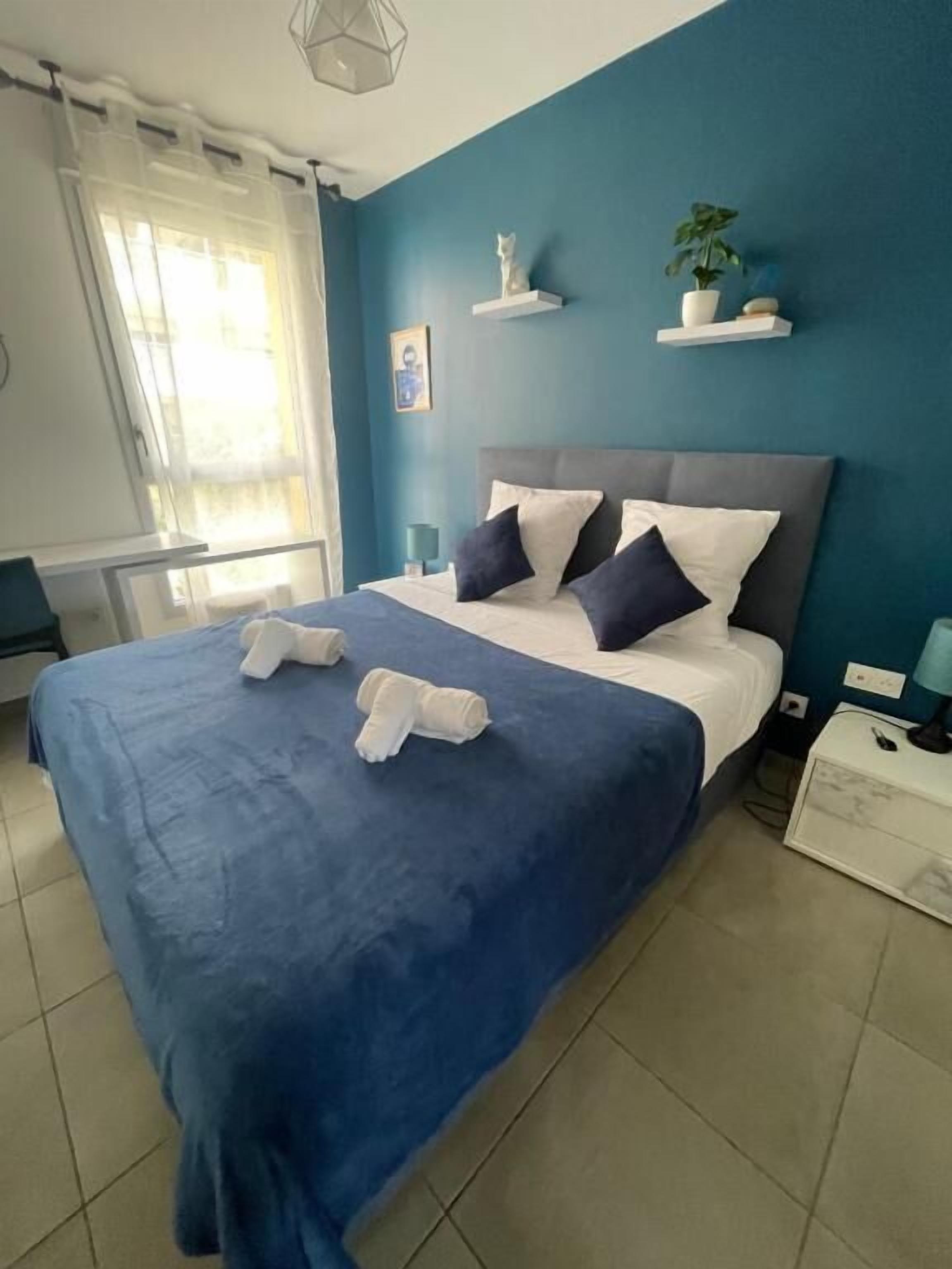 Camera da letto con un letto, cuscini, una coperta blu, un asciugamano bianco, un comodino, una lampada, una pianta in vaso e una mensola a muro con una piccola statua.