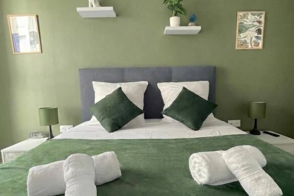 A neatly made bed with green and white bedding, two pillows, and two rolled towels on the bed.