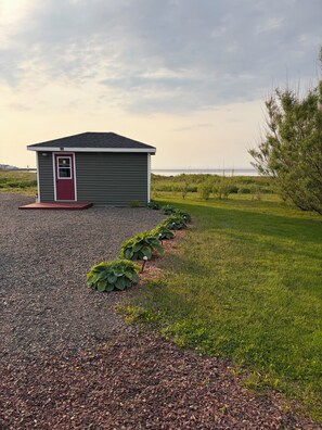 A small building with a red door, surrounded by gravel and grass, with a view of the water in the background. - Cozy home with a view of the ocean ! (Grand Bank)