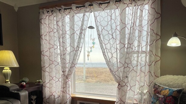 Ein Zimmer mit Holzboden, einem Fenster mit gemusterten Vorhängen, einer Lampe, einem Sofa und einem Sessel.