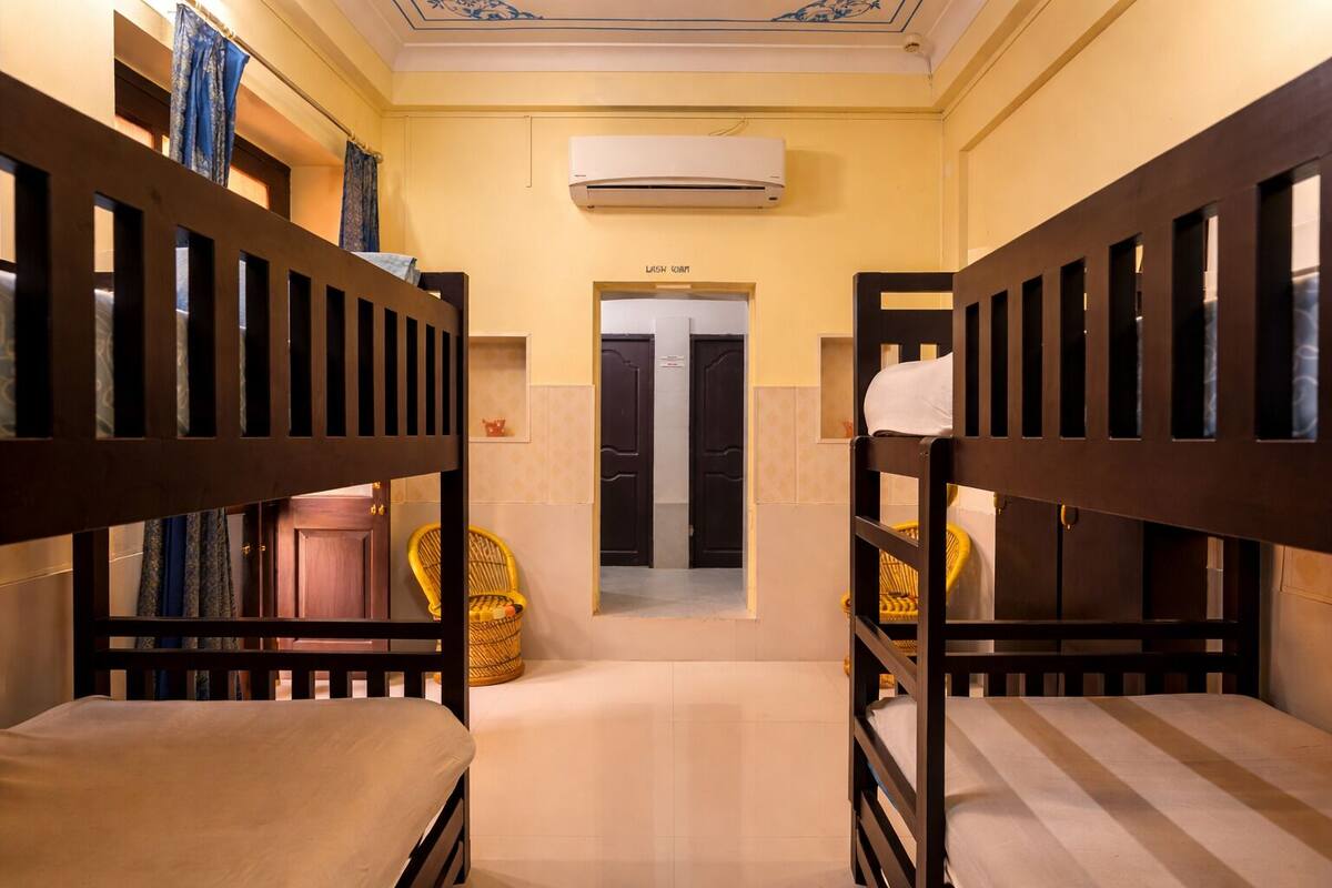 Ein Hotelzimmer mit Etagenbetten, einer kleinen Sitzecke mit einem Stuhl und einem Korb, einer Klimaanlage und einer Tür mit der Aufschrift 'LASH ROOM'.