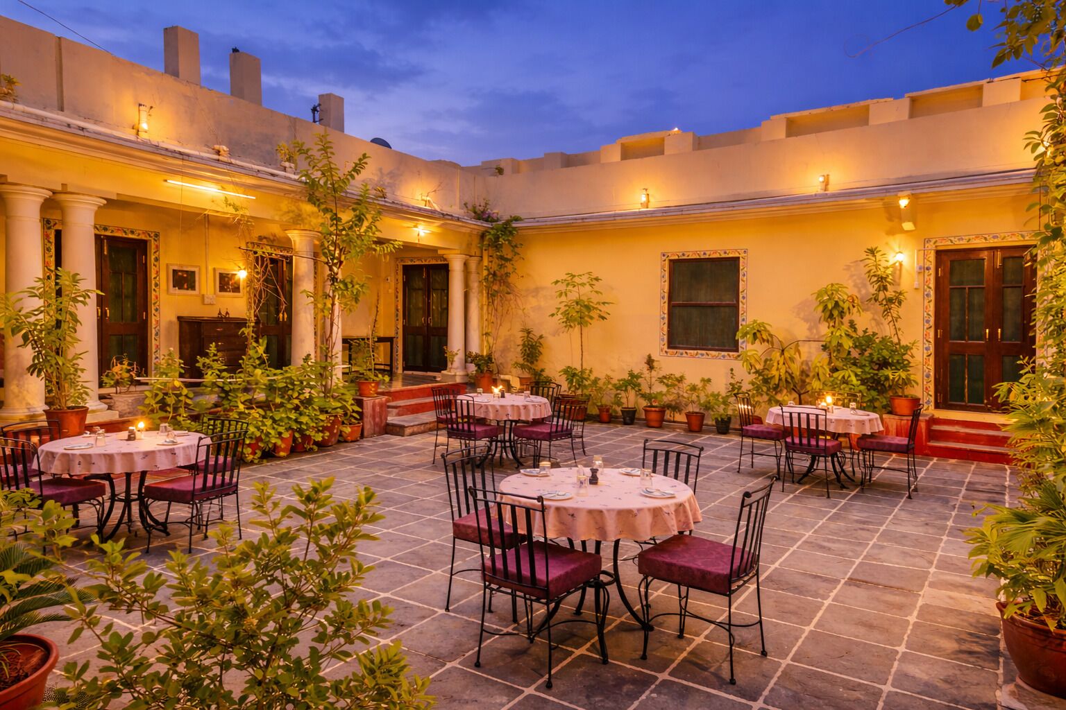 Un área de comedor al aire libre con mesas y sillas redondas, rodeada de macetas con plantas y columnas arquitectónicas.