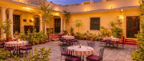 An outdoor dining area with round tables and chairs, surrounded by potted plants and architectural columns.
