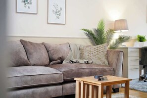Ein Wohnzimmer mit grauem Sofa, einem Holztisch, einer Lampe und gerahmten Pflanzenbildern an der Wand.