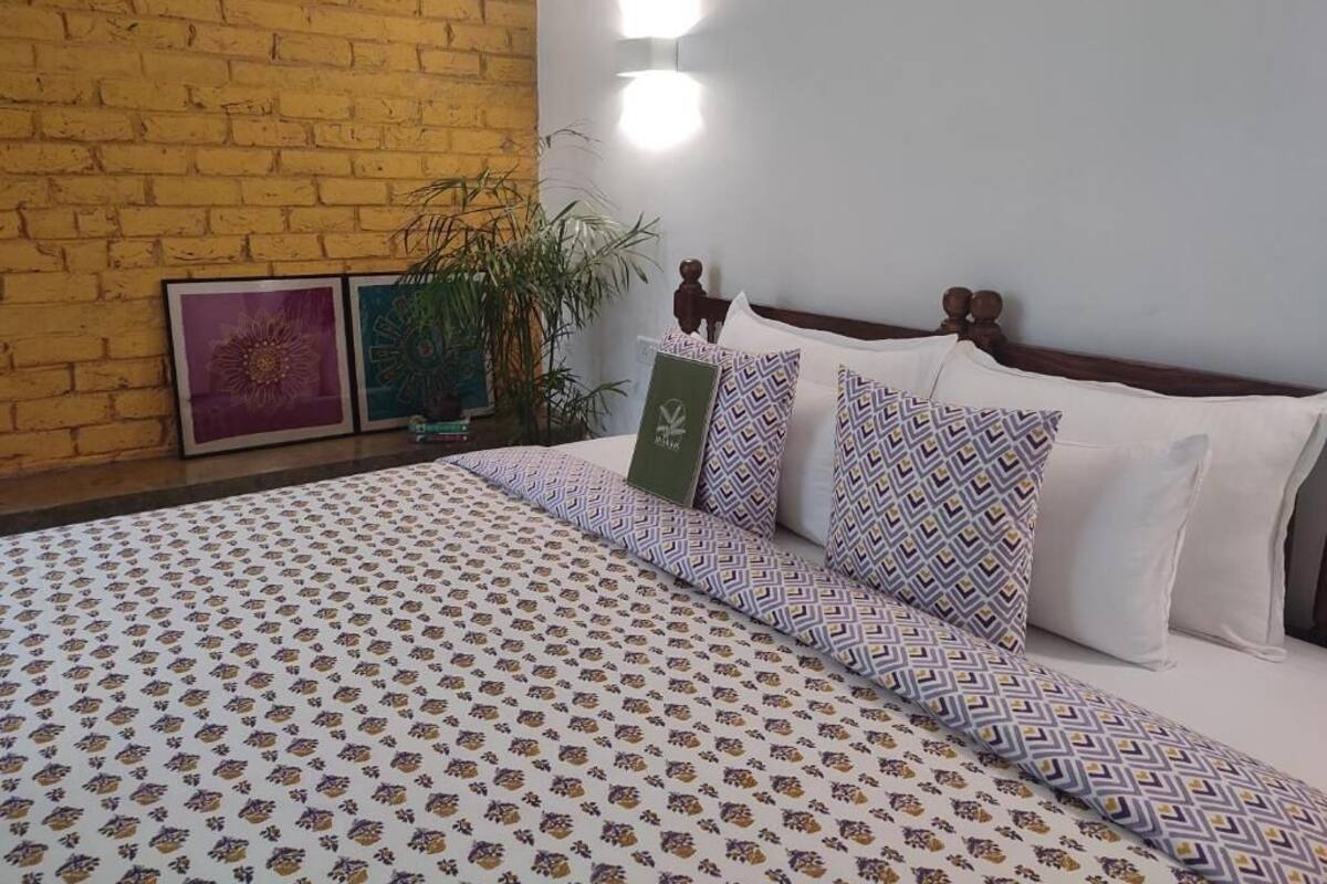 A hotel room with a brick wall, a bed with patterned bedding, a potted plant, and two wall-mounted lights.
