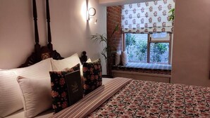 A bedroom with a bed, pillows, a bedside table, a nightstand, a window with a view, and a wall-mounted light fixture. - Mukam (Jaipur)