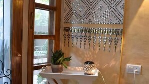 A room with a patterned floor, a white table with a plant, a decorative wall hanging, and a wicker chair. - Mukam (Jaipur)