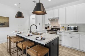 A modern kitchen with white cabinets, a central island with stools, a black pendant light, and stainless steel appliances. - Wayfarer's Loft at The Sojourn: Brand New 2BR,  Sophisticated Retreat, Downtown Ocean Springs, Pet Friendly, Golf Cart Rental (Ocean Springs)