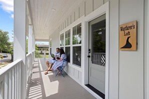 Un portico coperto con due persone sedute, un cartello di legno con la scritta 'Wayfarer's Loft' e una porta bianca con maniglia nera.