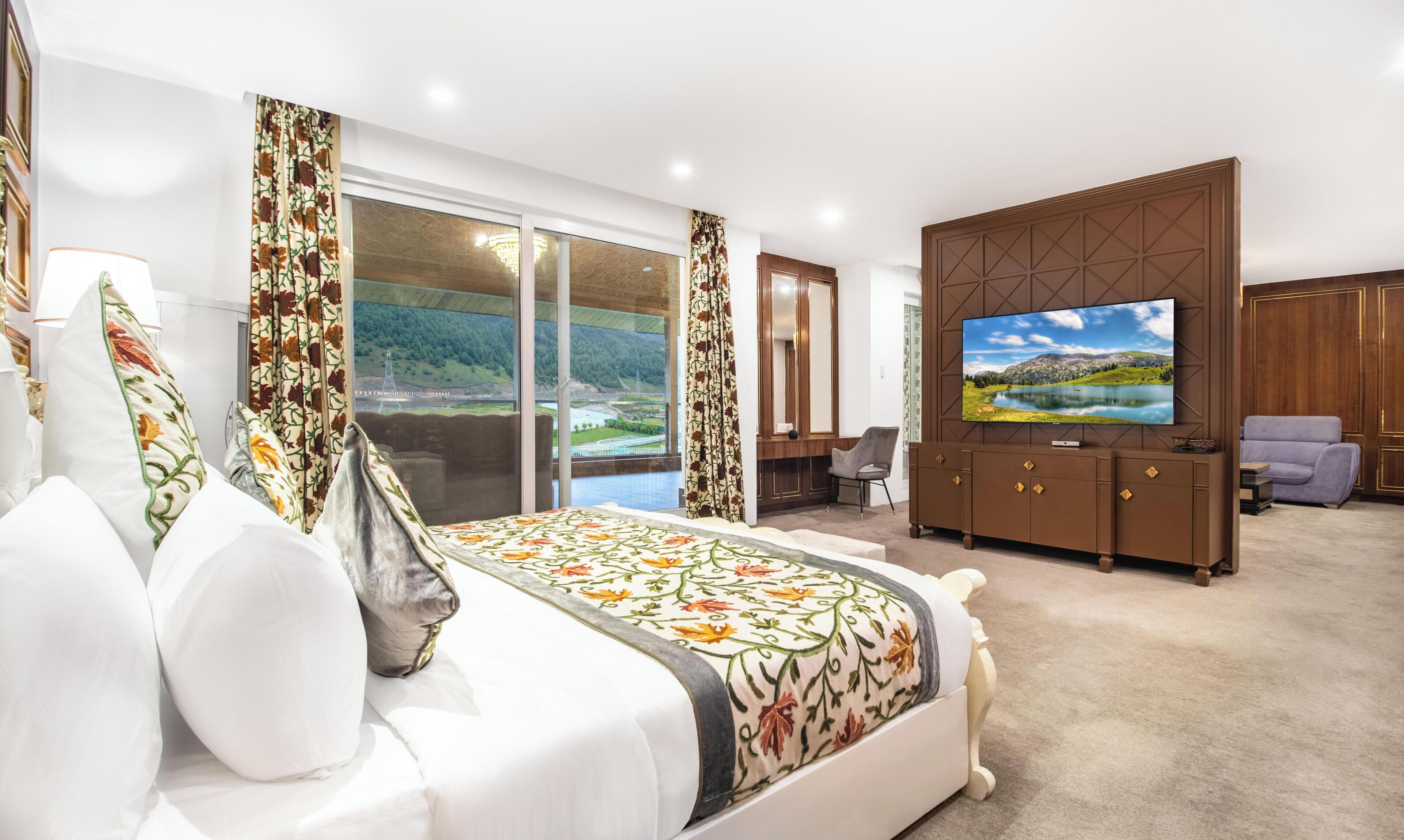 A hotel room with a bed, a television, a seating area, and a view of a mountainous landscape.