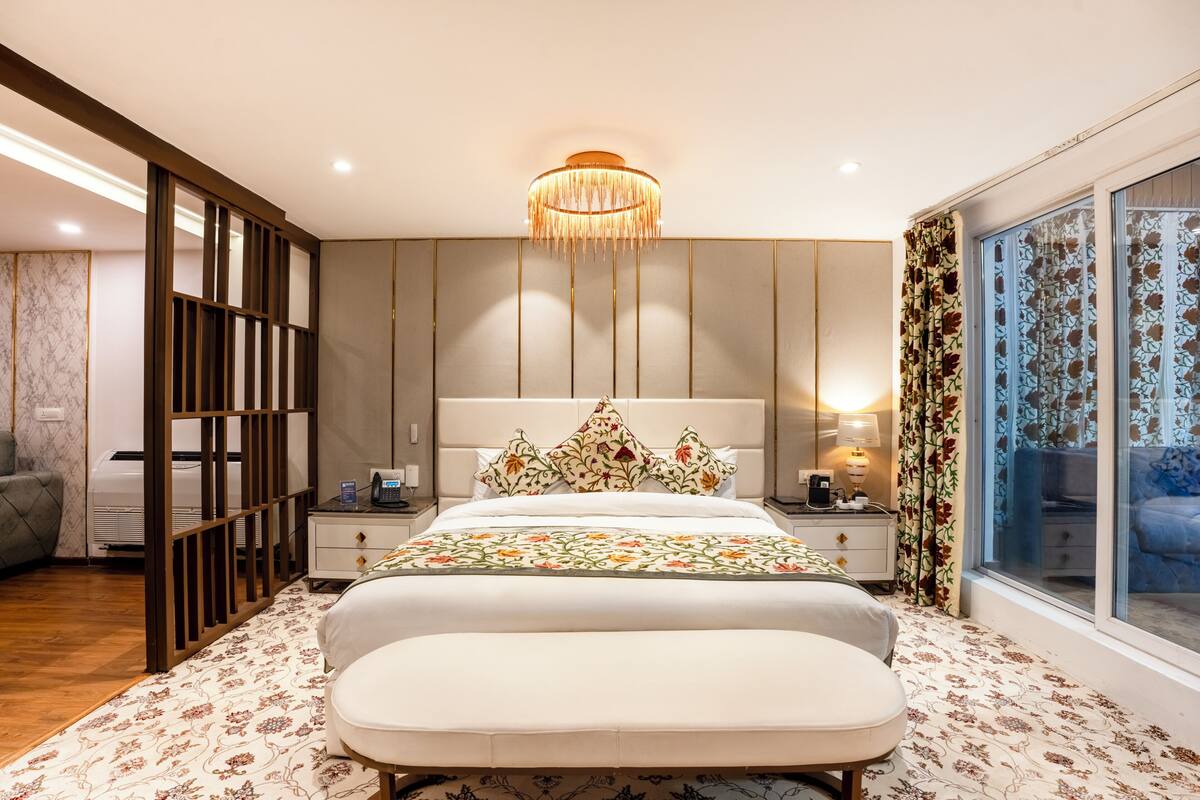 A bedroom with a bed, bedside tables, a nightstand, a decorative chandelier, a window with curtains, and a view of a balcony.