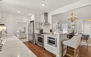 A modern kitchen with white cabinetry, stainless steel appliances, and a wooden dining area. - Hobnob - Private Pool - Golf Cart (Santa Rosa Beach)