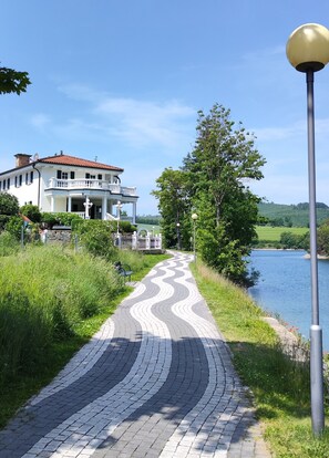 Ein Weg führt zu einem weißen Gebäude mit Balkon, umgeben von Grünpflanzen und einem Gewässer.