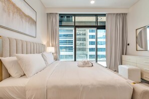 Ein Hotelzimmer mit einem großen Bett, einem Kopfteil, einem Nachttisch mit Lampe, einem Schminktisch und einem großen Fenster mit Vorhängen.