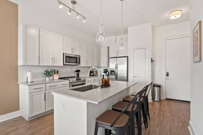 A modern kitchen with white cabinetry, stainless steel appliances, a central island with bar stools, pendant lighting, and a visible trash bin. - Skyline Suite - Free Parking, Pool, City Views (Charlotte)