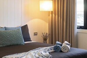 Una cama bien hecha con almohadas, una lámpara de noche, un florero con flores secas y una ventana con cortinas.