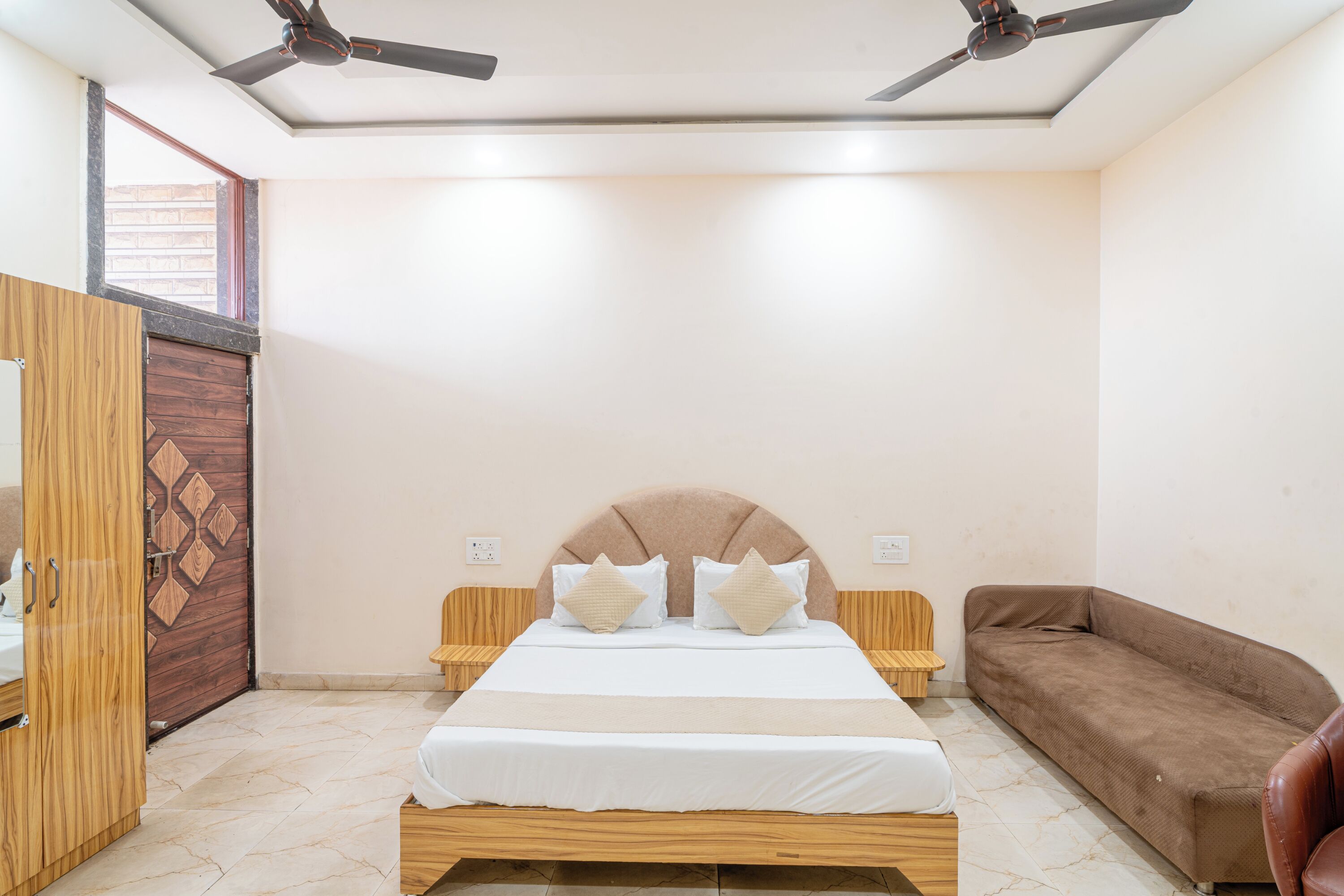 A bedroom with a bed, a brown sofa, wooden wardrobe, ceiling fans, and a window.