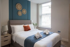 Cama bem arrumada com lençóis brancos, manta azul e toalhas dobradas. Há painéis de parede trançados decorativos acima da cama.