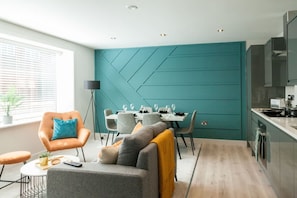Une cuisine moderne avec un mur dâaccent de couleur turquoise, un coin repas avec une table blanche et des chaises grises, et un coin salon confortable avec un canapĂ© gris et un fauteuil orange.