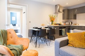 Een moderne woonkamer met een bank, een eettafel met stoelen, een keuken met kasten en apparatuur, en een doorgang naar een andere kamer.