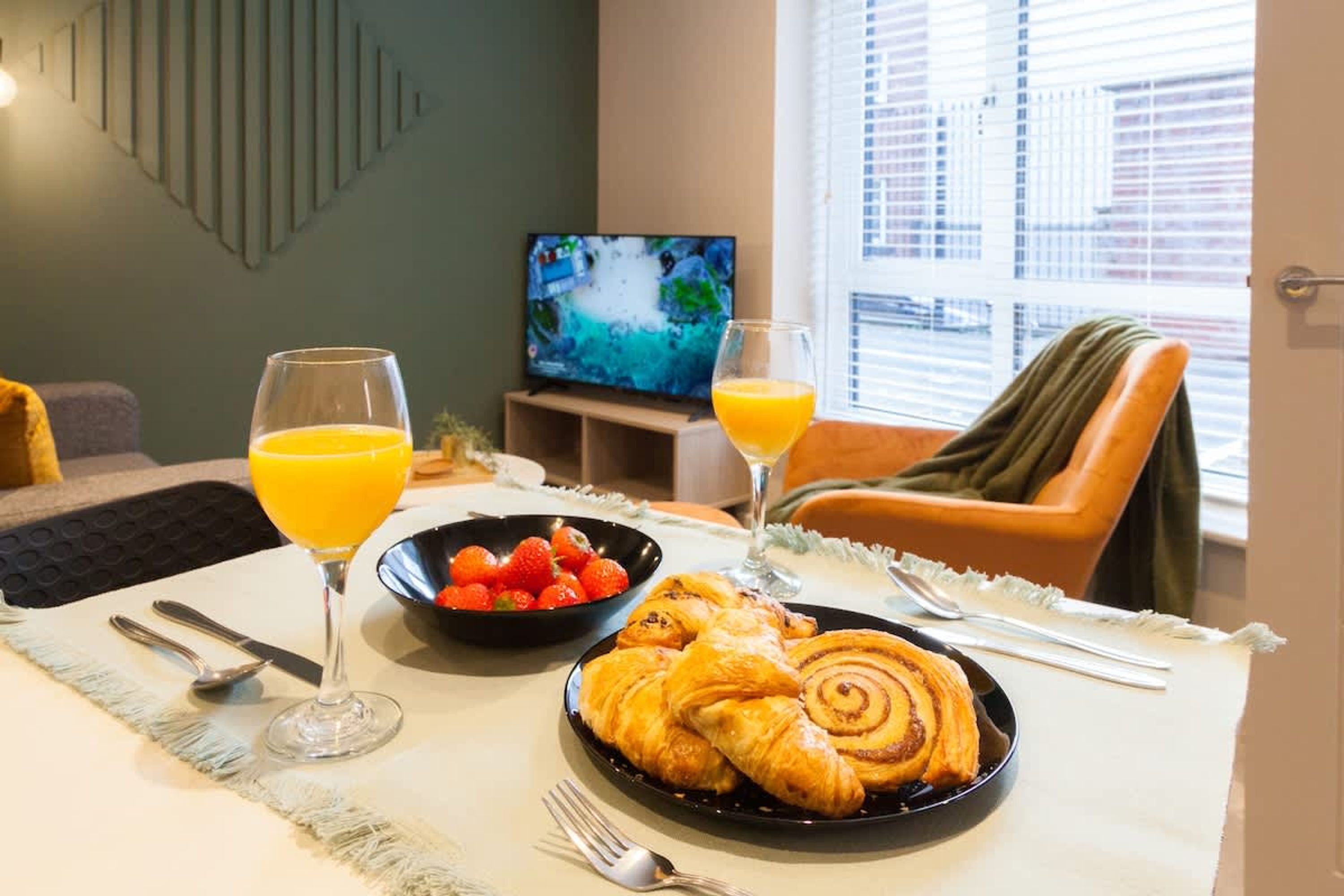 A breakfast setup with croissants, strawberries, and orange juice on a table, with a TV in the background.
