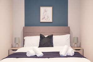 Een net opgemaakt bed met witte en donkere kussens, een lijstwerk erboven en twee bedlampen aan weerszijden.