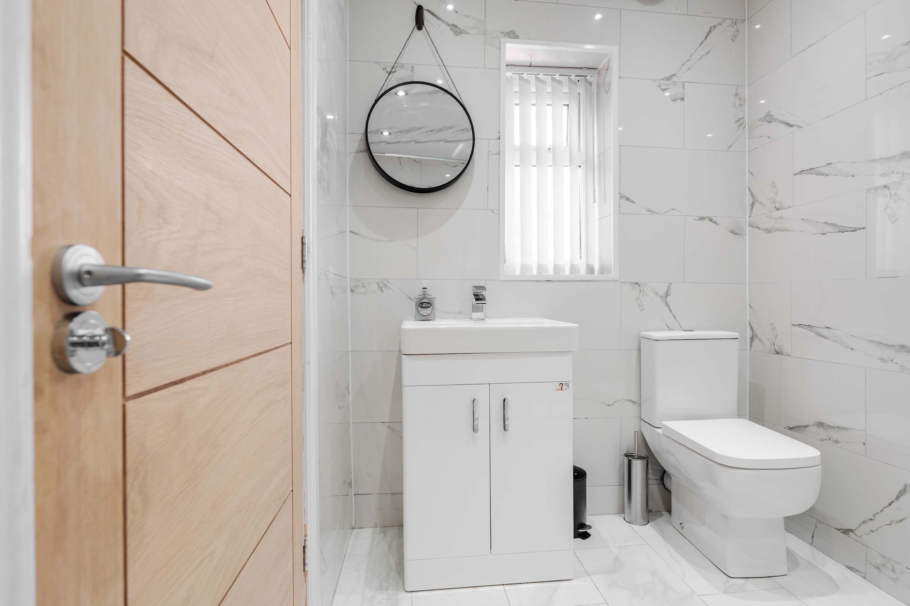 A modern bathroom with a wooden door, a white sink, a toilet, and a marble-tiled wall.