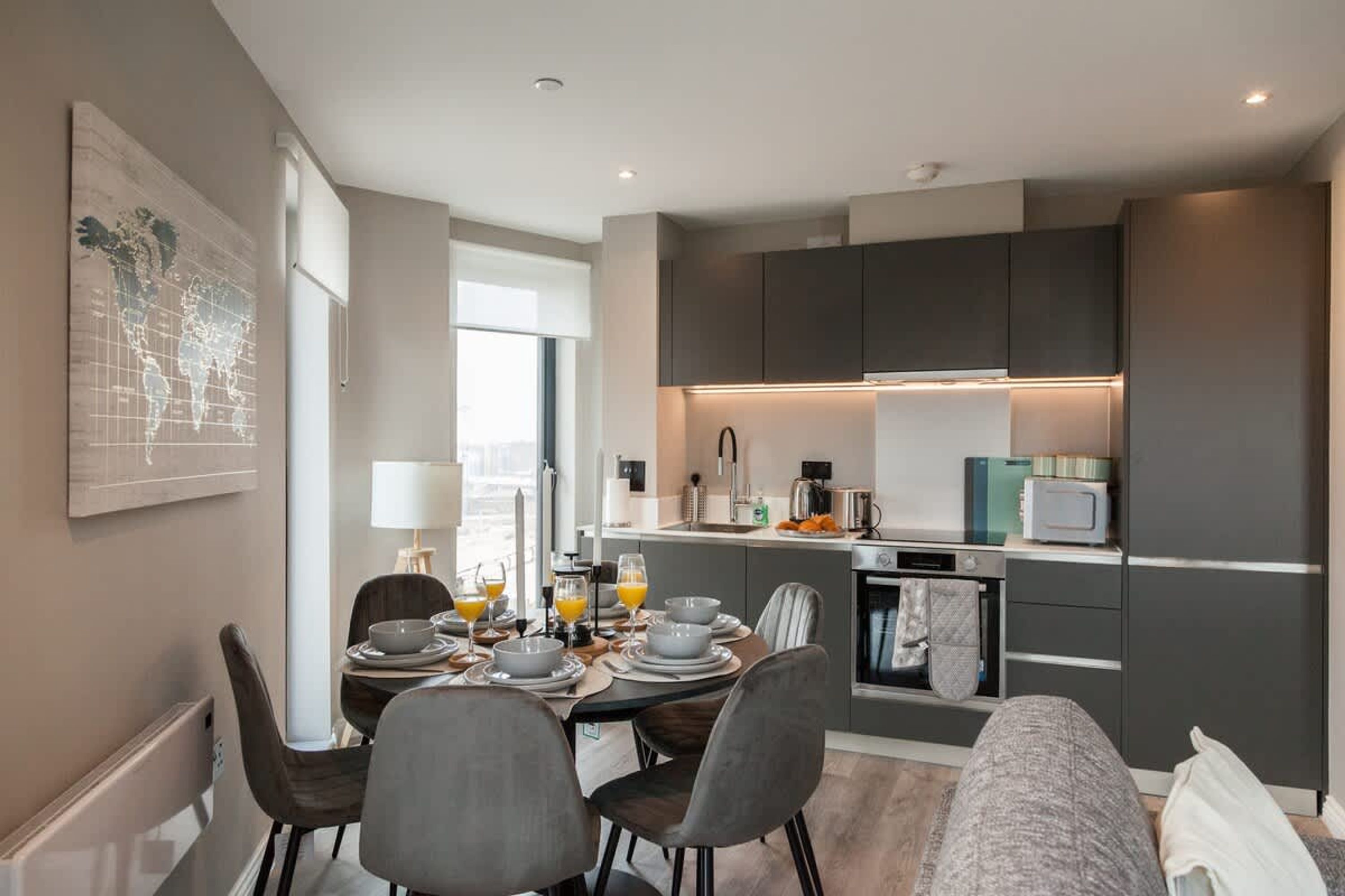 Una cocina moderna con gabinetes oscuros, un comedor con una mesa dispuesta para cuatro personas y un ventanal que proporciona luz natural.